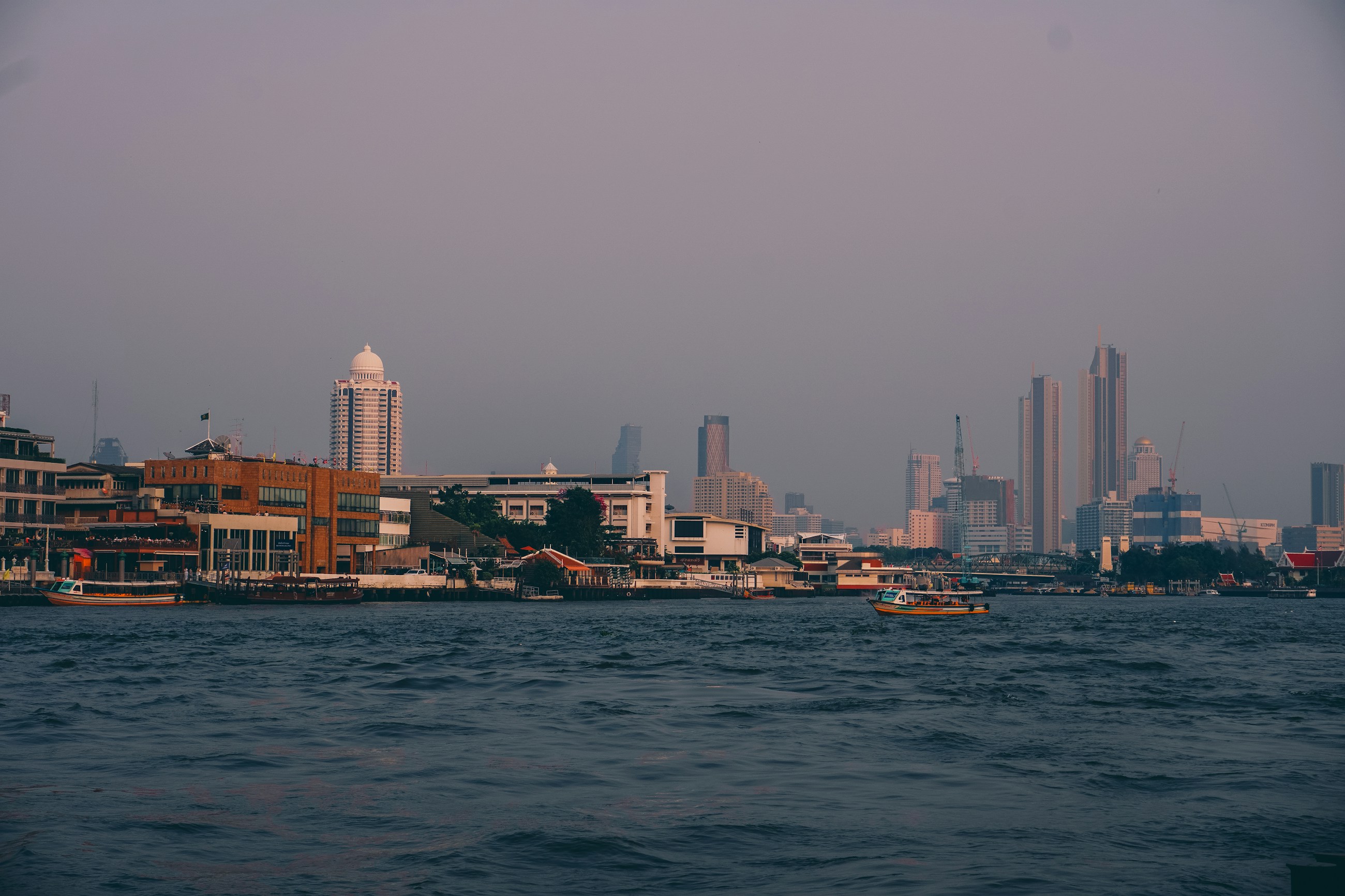Bangkok skyline and Chao Phraya river boats at dusk for a Riverside versus Sukhumvit hotel guide.